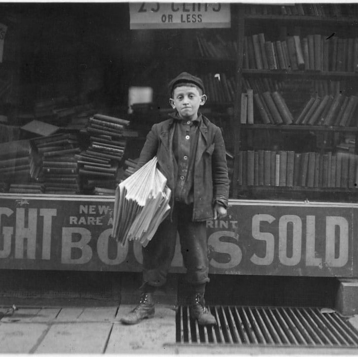 Young boy in a cap and coat stands outside a newspaper shop, holding a large stack of folded papers for sale.