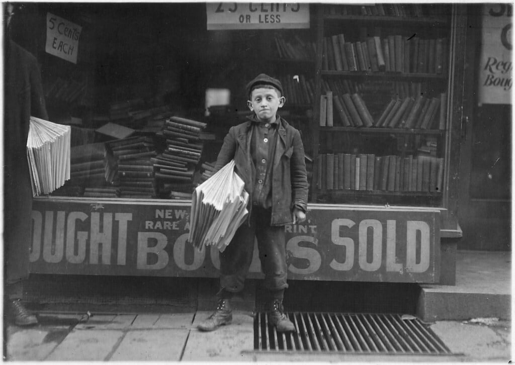 An old black and white photo of a newsboy holding perhaps 20 newspapers standing in front of a newsstand.
