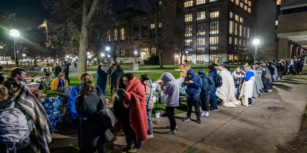 Long line of people wrapped in blankets outside a campus building at night waiting to vote.