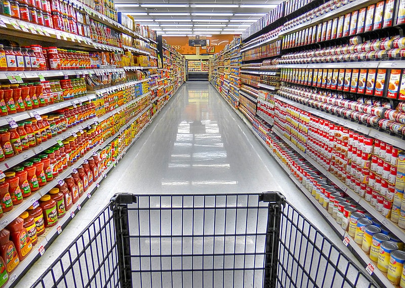 Shopping cart in a supermarket aisle with tall shelves stocked with colorful condiments on both sides.